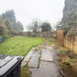 Overgrown garden with stone wall and bins.